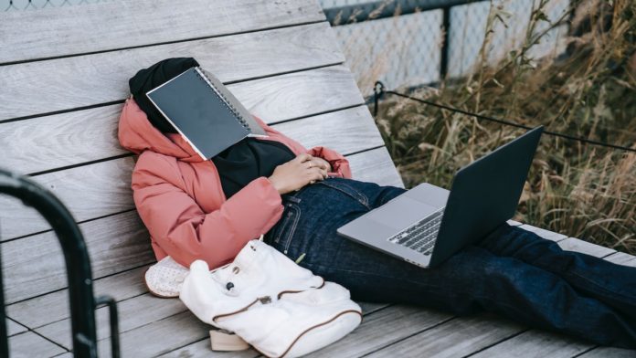 Person laying on bench with laptop on his knees and book on his head dreaming