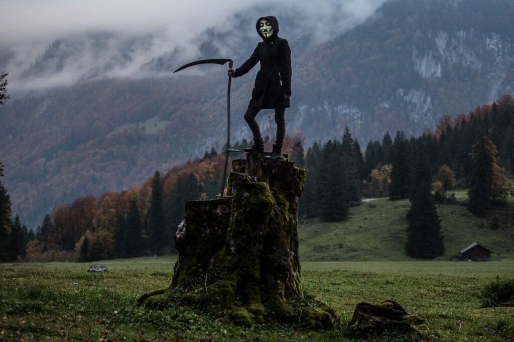 A man in anonymous vendetta mask stands on a cut tree with a sickle